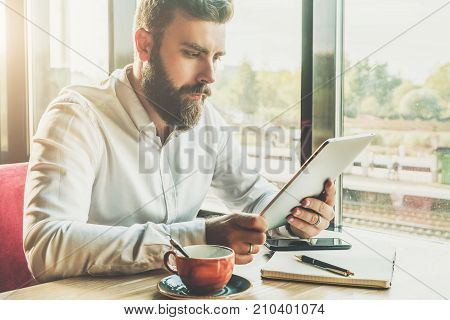 Young bearded businessman sits in cafe at table, holding tablet computer, working.On table cup of coffee