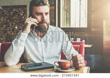 Young bearded businessman sits in cafe at table, drinking tea, talking on cellphone. On table notebook, pen. Man is working
