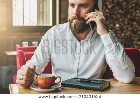 Young bearded businessman sits in cafe at table, drinking tea, talking on cellphone. On table notebook, pen. Man is working