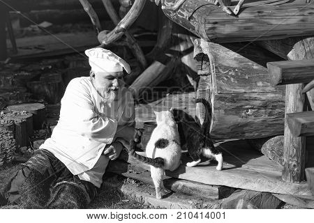 bearded man cook chef in uniform and hat with long beard on smiling face holding iron old tea kettle near black and white cats sunny day outdoor on wooden background