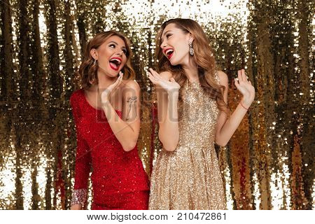 Portrait of two cheery beautiful women in sparkly dresses looking at each oher while standing and laughing together isolated over golden shiny background