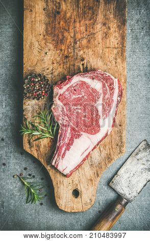 Flat-lay of raw prime beef meat dry-aged steak rib-eye on bone with seasoning and chopper on rustic wooden board over grey concrete background, top view, copy space. Meat high-protein dinner concept