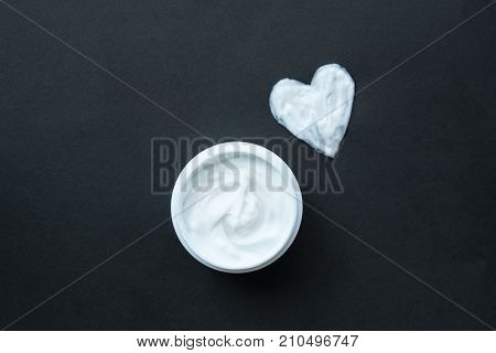 Cream for face or body in white container and femile hand. Heart shape from cream on black background
