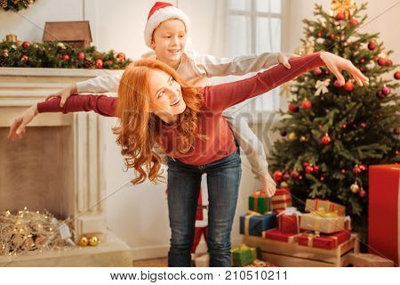 No time for worries. Positive minded mother and son grinning broadly while pretending to fly and playing together on a Christmas morning.