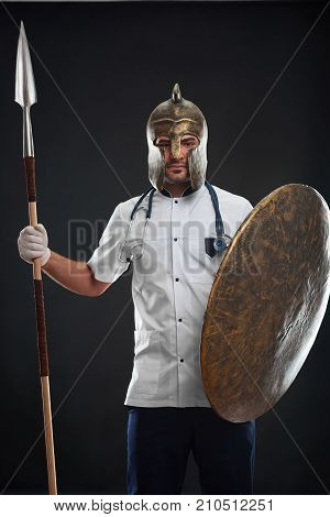 Vertical shot of a young handsome male doctor wearing a helmet posing with a shield and a spear on black background at studio health medicine protection fighting disease vitality concept.