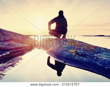 Man Sit At Evening Sea. Hiker Sit In Squatting Position On Granite Cliff Above Smooth Water.