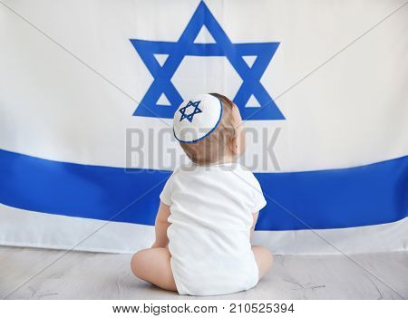 Cute baby in kippah sitting on floor against big flag of Israel