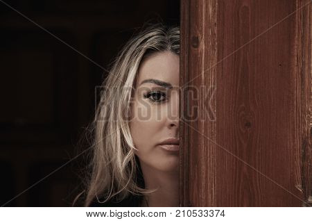 Beautiful girl peeks through the door peeking Out of Doorway. GIrl with low lighting a massive door covers half face closeup. Suspicious and attentive look.