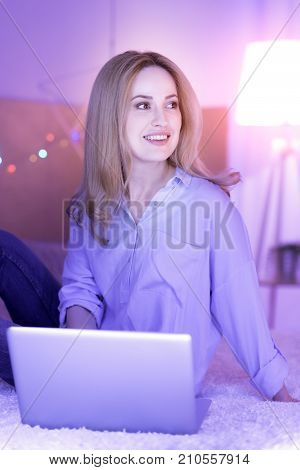 My dream work. Good-looking exuberant young lady sitting on the sofa and smiling while working on the laptop and looking somewhere