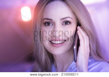 Happy eyes. Beautiful dark-eyed blond woman smiling and feeling on cloud nine while talking on the phone