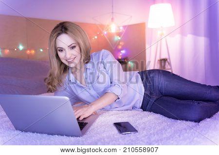 Businesswoman. Sweet cheerful young woman smiling while working on her laptop and lying in bed with her phone by her side