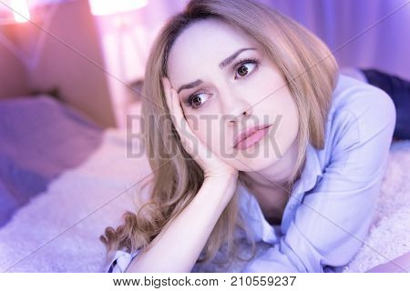 In the blues. Good-looking young blond woman lying on bed holding her head with her hand and looking sad