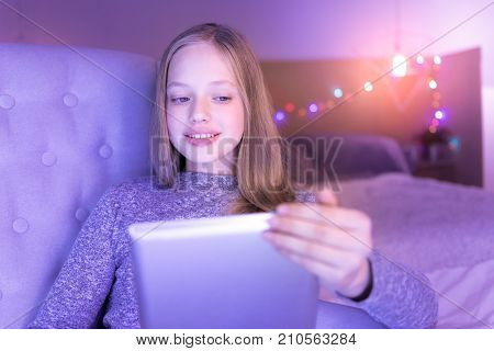 Watching photos. Delighted blue-eyed blond girl smiling while sitting and looking at her tablet