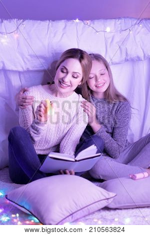 Enjoying reading. Alive young loving mother and daughter hugging and mom holding a torch while reading a book