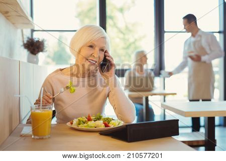 Good news. Happy cheerful senior woman smiling and making a call while enjoying her lunch