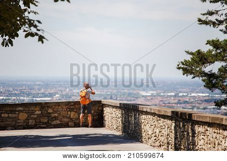 The Tourist Man Stands With His Back On The Viewing Platform In Bergamo In Italy And Takes A Photo O