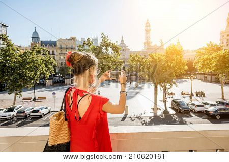 Young woman tourist in red dress photographing Ayuntamiento square from the terrace of city hall building in Valencia during the morning light in Spain