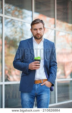 Attractive business man with beard drinking coffee and posing on the camera on the street. Panoramic wondows on the background. Close-up of man.