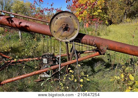 An old rusty tube grain elevator with motor and cable lift wheel is left in an autumn colored grove.