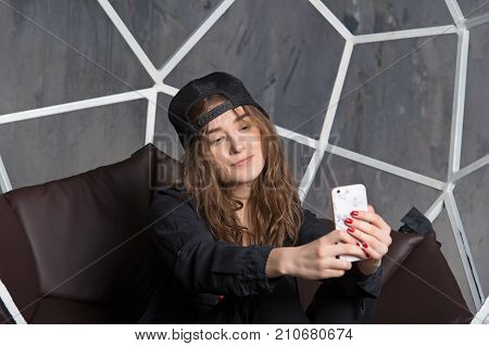 Girl or woman in baseball cap making selfie with smartphone on brown chair. Technology and mobile device. Self expression concept.