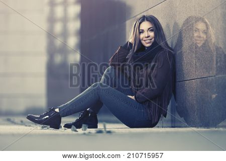 Happy young woman sitting at the wall in city street. Stylish fashion model in leather fringe suede jacket and blue jeans