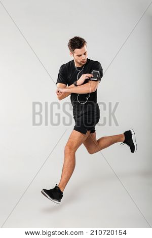 Full length portrait of a handsome young man athlete in earphones listening to music while running and touching his phone in armband isolated over gray background