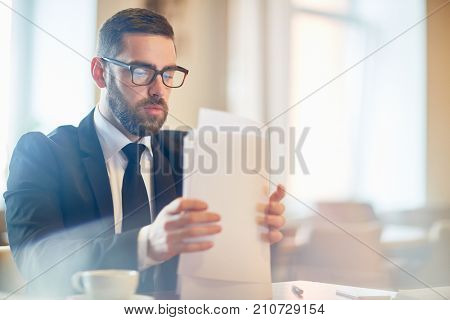 Confident bearded manager in classical suit sitting at office desk and doing paperwork, waist-up portrait
