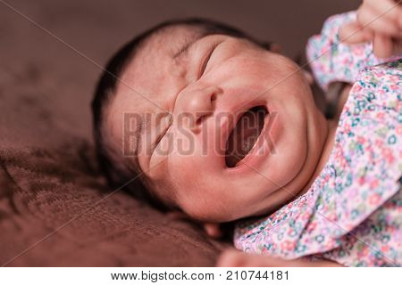Close up portrait of an unhappy cute two weeks old newborn baby girl lying down and crying hard, with cramps or colic, bellyache, stomach pain