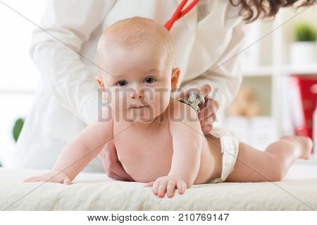 Pediatrician examines newborn child. Doctor using a stethoscope to listen to baby's back checking heart beat. Kid is looking at camera.