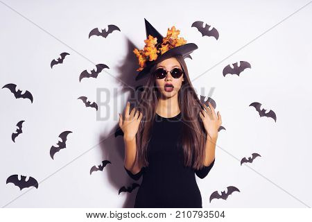 a fashionable Helluin witch in a big hat with leaves in round sun-protective glasses looks at the camera. the girl is going to the Halloween party. bats on white background