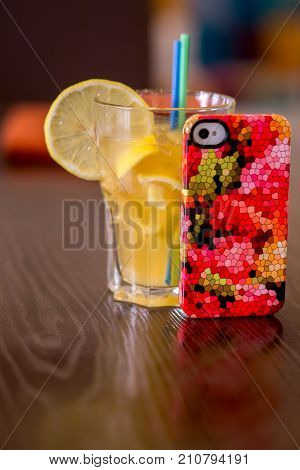 A bright multi-colored cell-phone cover on a table next to a juice coctail in a glass with a drinking straw and an orange slice.