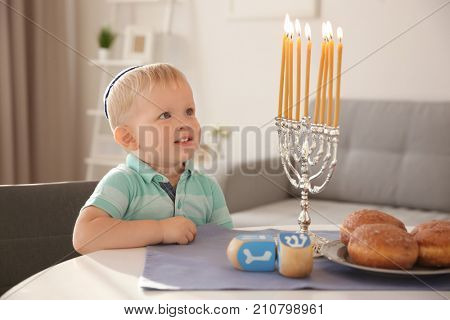 Cute boy sitting near nine-branched menorah on table at home