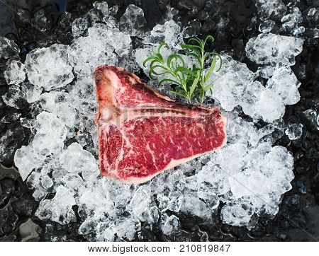 Raw fresh meat t-bone steak on chipped ice with rosemary over dark slate stone backdrop, top view