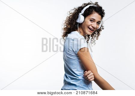 Mood-boosting music. Charming upbeat curly-haired woman listening to the music in headphones and laughing while standing half-turned against a white background