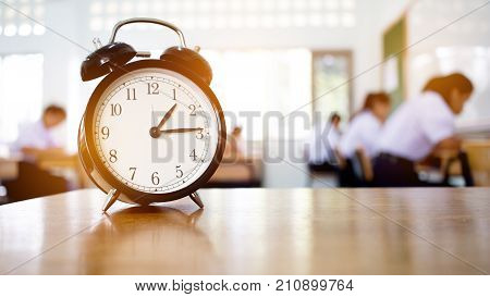 Retro alarm clock with one o'clock left side on wood table teacher in blur students exams classroom. Time is indefinite continued progress of existence. Education exams concept black and white selective focus.