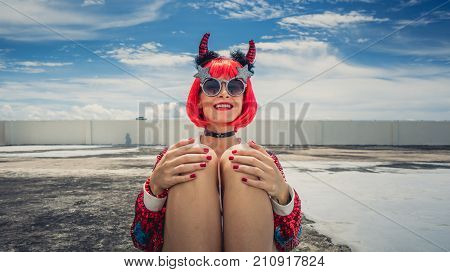 Halloween. Portrait of a beautiful girl in a red wig with horns close-up against a blue sky. Sitting on the foof. Red, holiday makeup