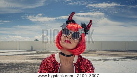 Halloween. Portrait of a beautiful girl in a red wig with horns close-up against a blue sky. Sitting on the foof. Red, holiday makeup