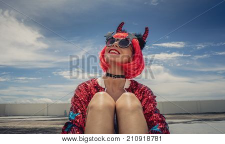 Halloween. Portrait of a beautiful girl in a red wig with horns close-up against a blue sky. Sitting on the foof. Red, holiday makeup
