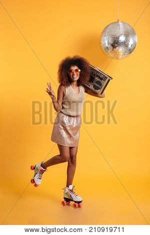Full length portrait of smiling afro american disco woman in sunglasses showing peace gesture, standing on roller skates, holding boombox, looking at camera, isolated on yellow background