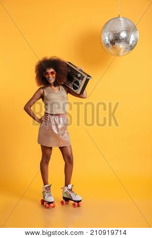 Full length portrait of cheerful african disco woman with hand on her waist wearing in retro clothes standing on roller skates, holding boombox, looking at camera, isolated on yellow background