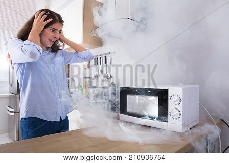 Unhappy Young Woman Looking At Smoke Emitting Through Microwave Oven In Kitchen