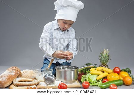 Cuisine Concepts. Portrait of Fascinated Caucasian Little Boy Crashing Fresh Egg In Cooking Hat. Surrounded by Fresh Fruits and Vegetables In Studio. Posing with Smiling Expression. Against Gray. Horizontal Image Orientation