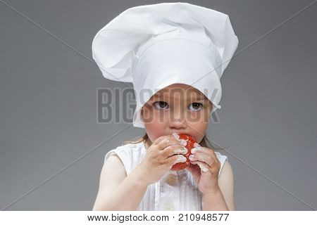 Cooking Ideas. Portrait of Funny Caucasian Little Girl in Cook Uniform Posing Fresh Tomato and with Smudgy Face Against Gray Background. Horizontal Image Orientation