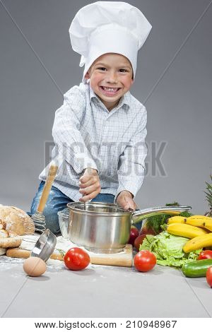 Portrait of Positive Smiling Caucasian Boy Working With Kitchen Ware In Cooking Hat. Surrounded by Fresh Fruits and Vegetables In Studio. Posing with Smiling Expression. Against Gray.Vertical Image