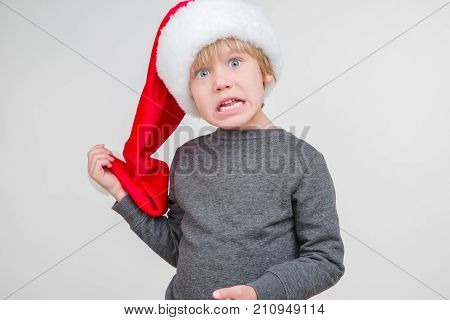 Little blond emotional boy in a festive red hat in a gray jacket. Little Santa Claus on a gray background.