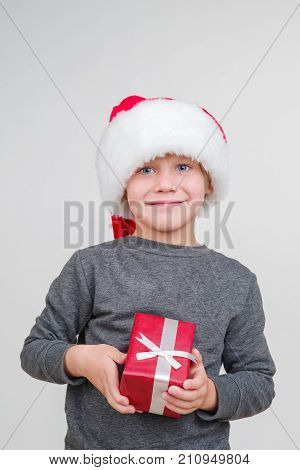 Little blond emotional boy in a festive red hat in a gray sweater with gifts. Little Santa Claus.
