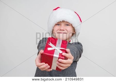 Little blond emotional boy in a festive red hat in a gray sweater with gifts. Little Santa Claus on a gray background.
