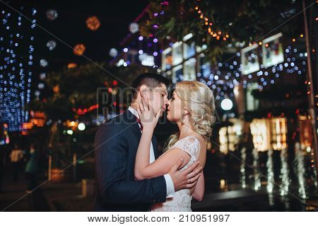 A couple of newlyweds kiss gently on the street in the evening surrounded by city lights.