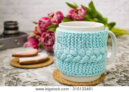 A White Cup Of Tea In A Knitted Blue Case With An Old Camera And Pink Flowers Tulips And Sweets, Gin