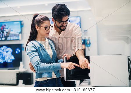 Young Modern Attractive Couple In The Tech Store Deciding Which Microwave To Buy.